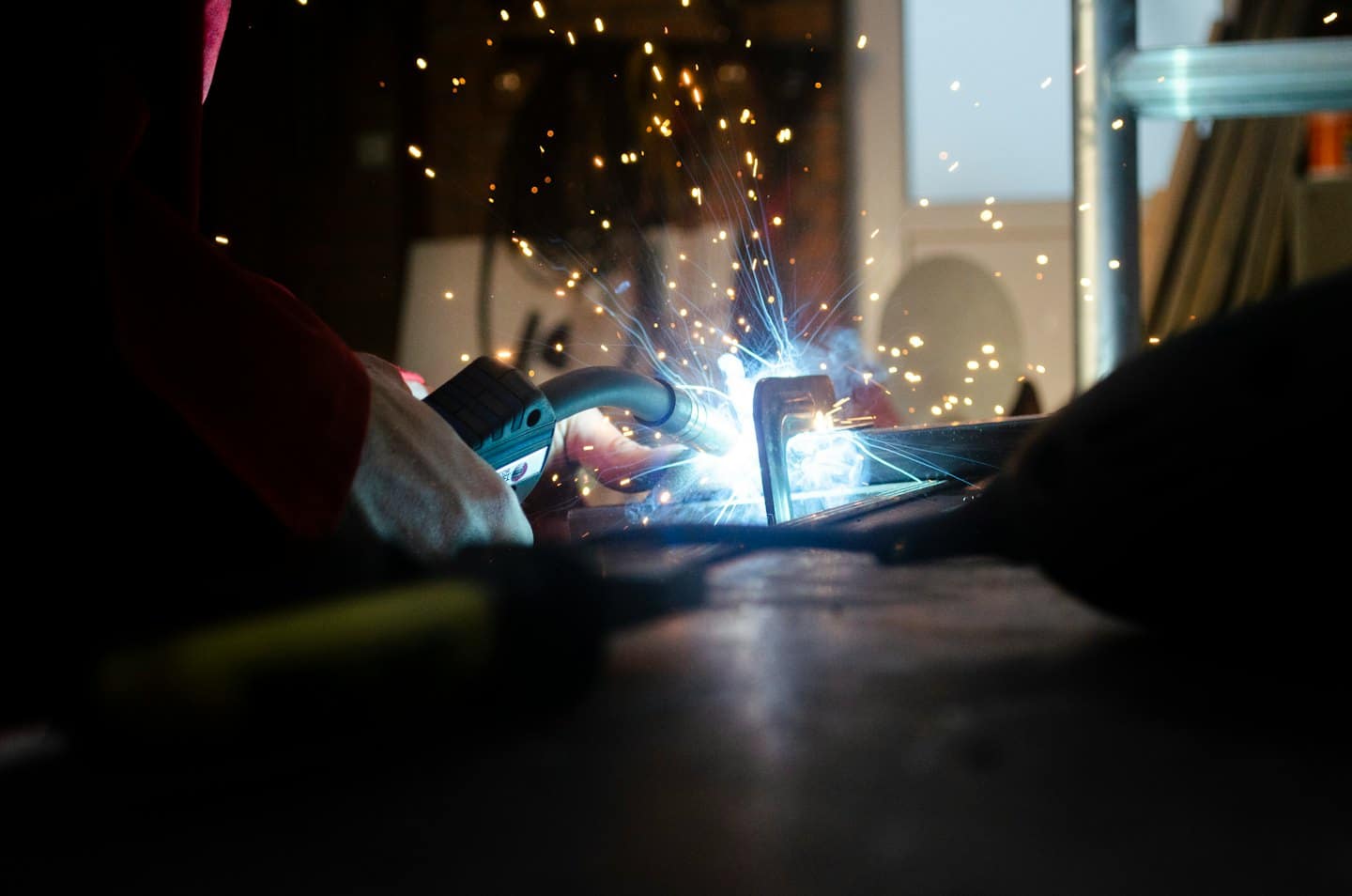 Hands in red sleeve MIG welding, sparks flying.